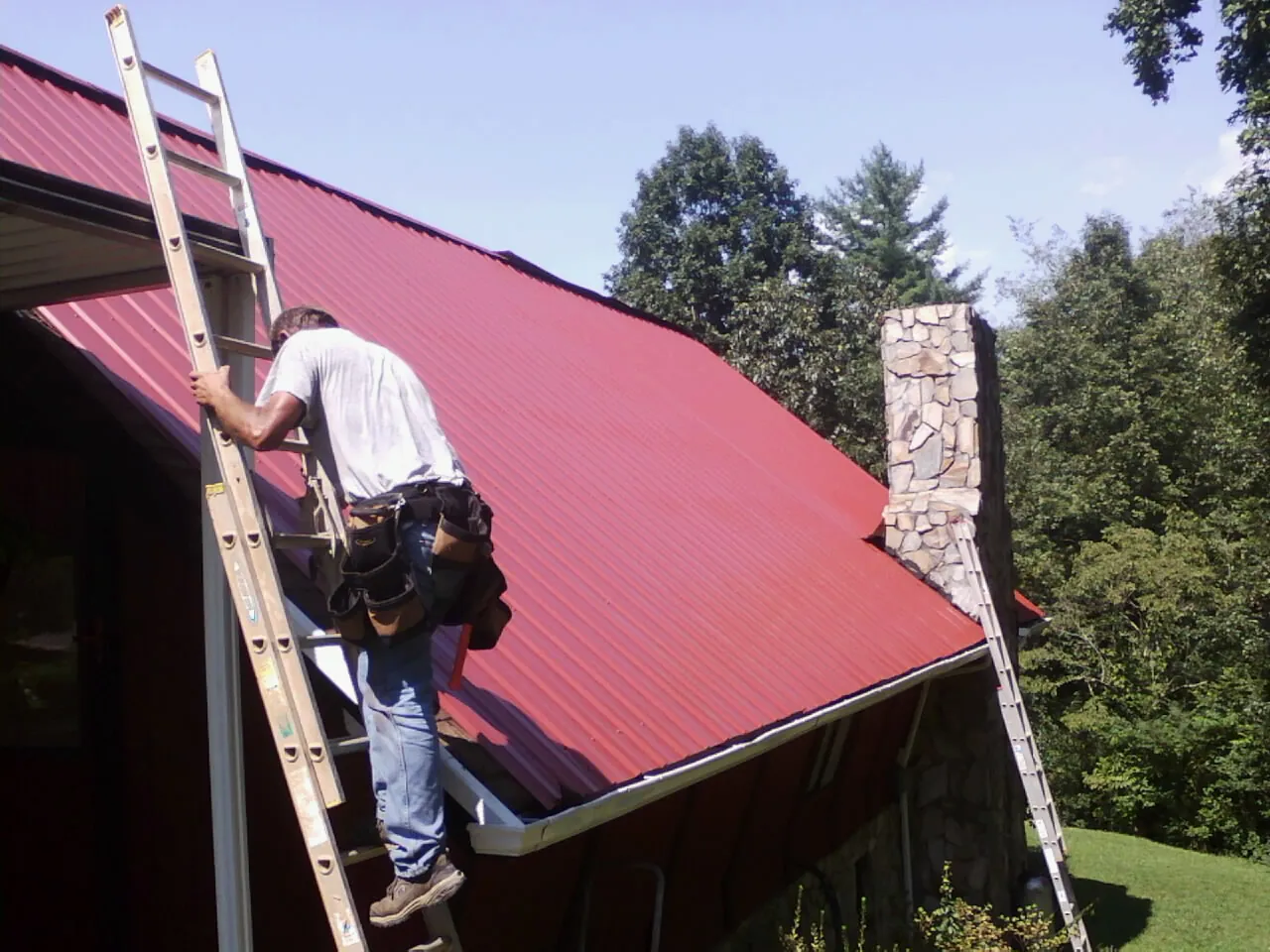 Roofer on a ladder beside a red metal roof and stone chimney — Soffit & Fascia Repair by Roofing Brothers in Pleasant Prairie